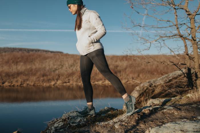 a woman in a white jumper and LIVSN trail tights hiking on the shore of a lake