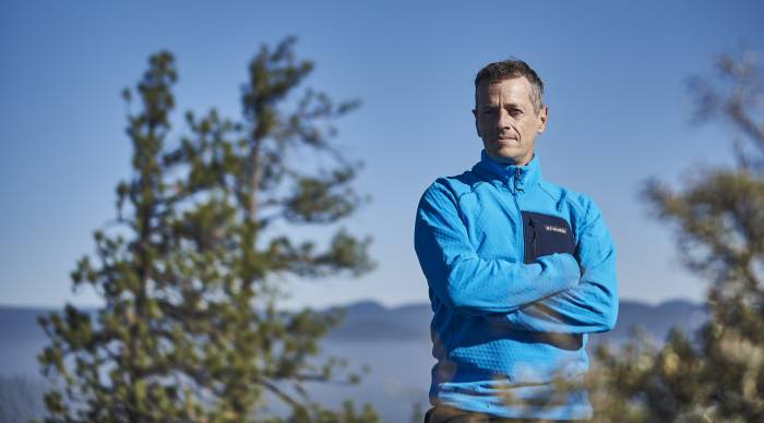 Race director and adventure racing pro Jason Magness stands on top of a mountain with clouds and trees in the background.