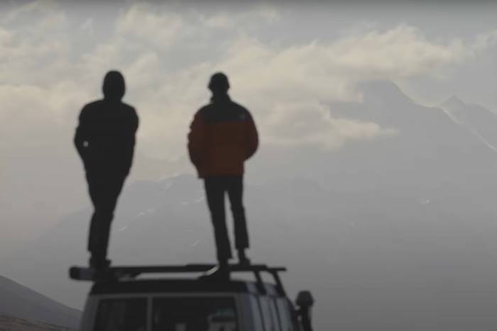 Wwo people stand on the roof of a van gazing at mountains