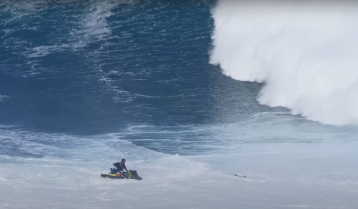 a surfer gets a tow from a jetski as a big wave looms behind