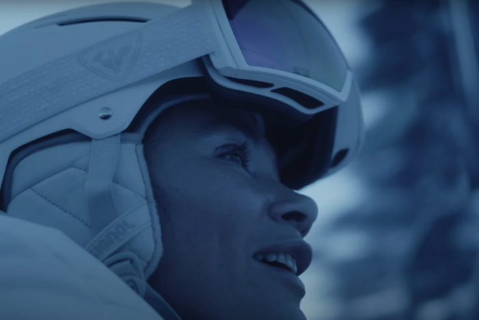 A woman wearing a ski helmet and goggles rides a ski lift in morning light
