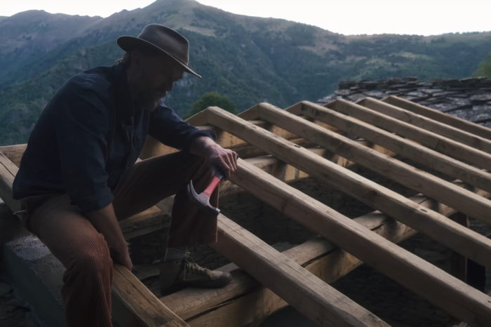 A man holding a hammer sits atop a partially constructed roof with mountains in the background