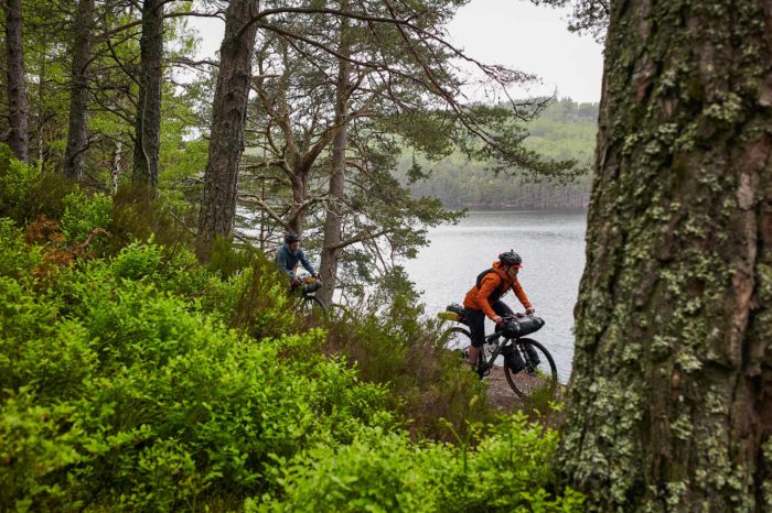 Two people on a bikepacking trip ride a trail near a lake.