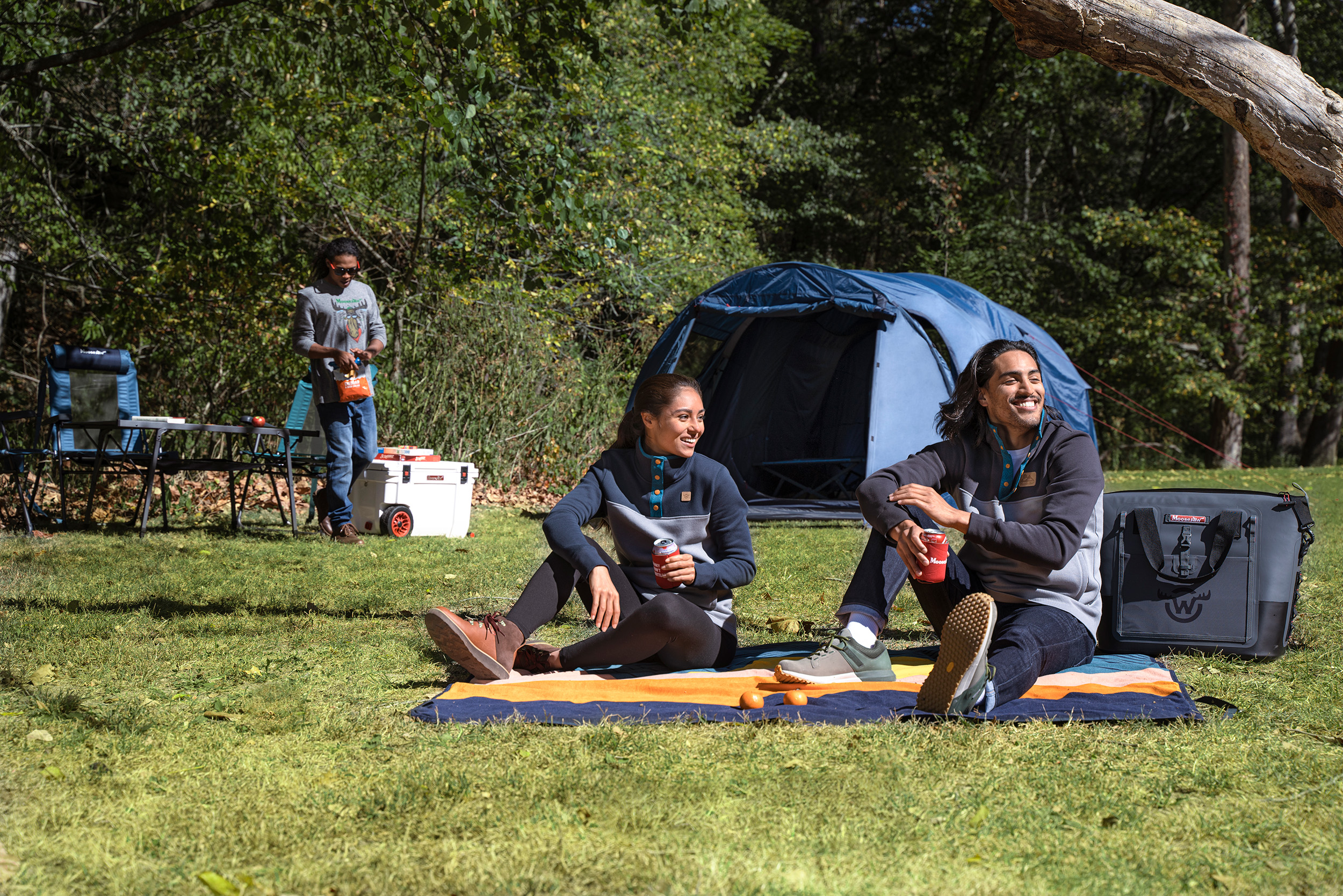 Two people sitting on a camp blanket with a tent in the background.