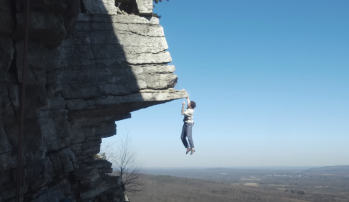 free solo climber hangs off a tall rock prow with one arm