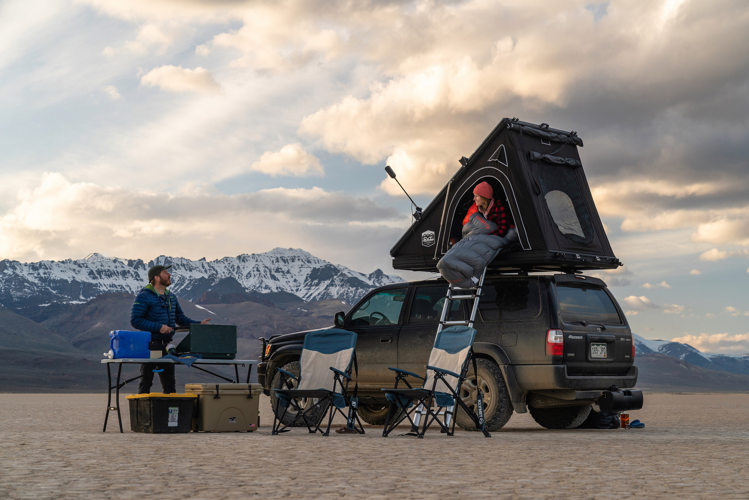 Two people camped in a desert with a vehicle mounted shelter and a slew of camping gear.