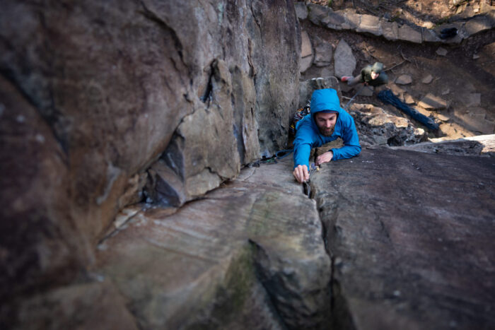 Man rock climbs while wearing a windbreaker jacket and placing protection