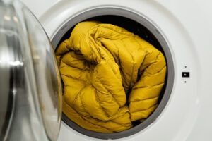 Yellow winter jacket in the drum of open washing machine in laundry room. Washing dirty down jacket in the washer
