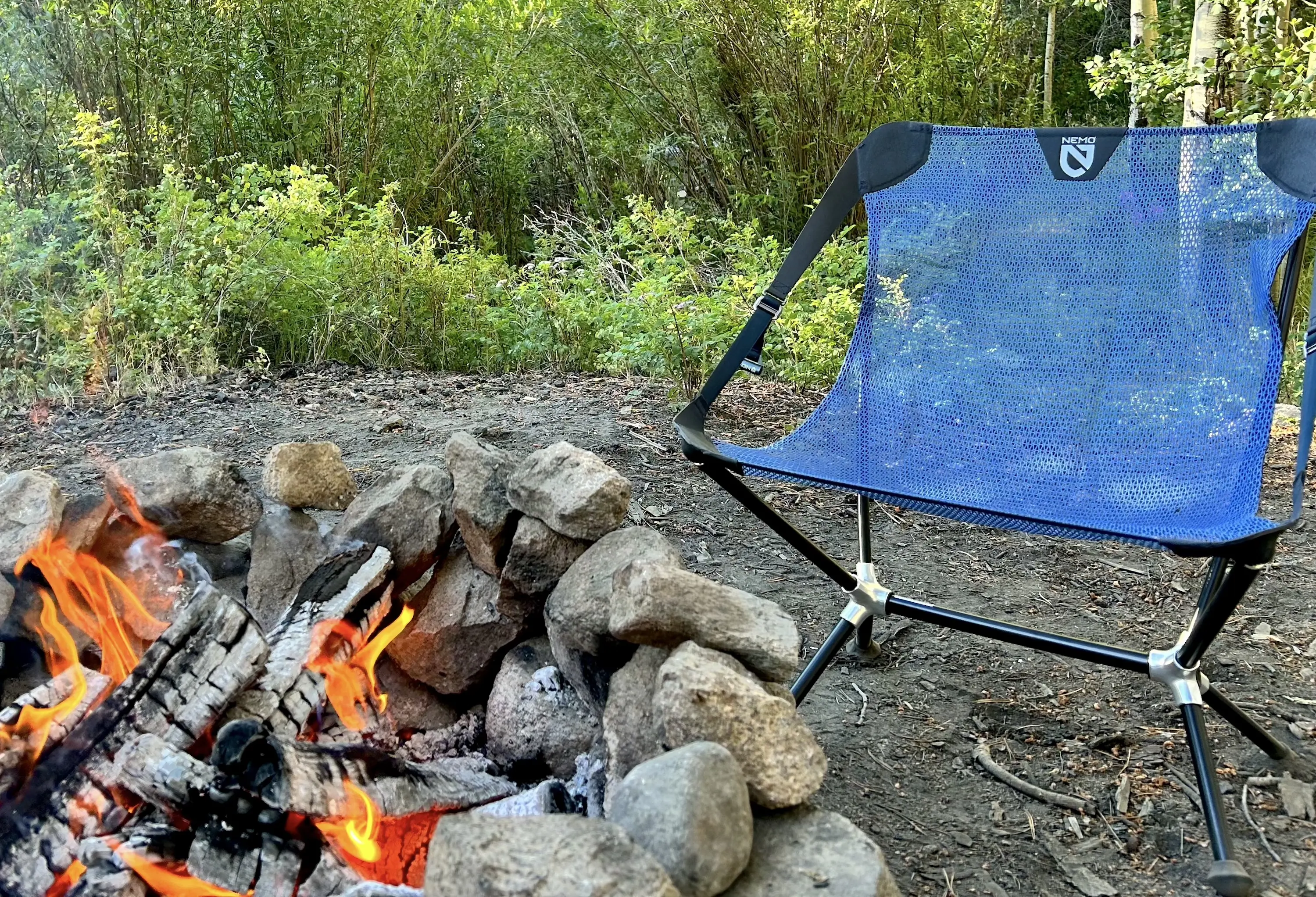 a blue mesh NEMO backpacking chair next to a big rock ring with a campfire during the summer