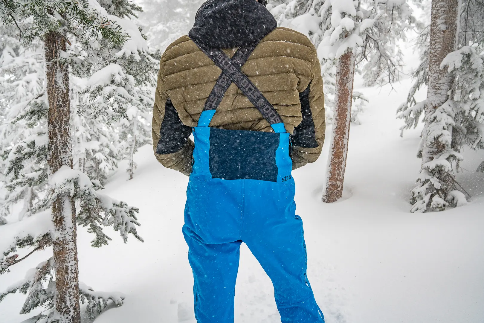 man wearing down jacket beneath ski bibs during snow storm 