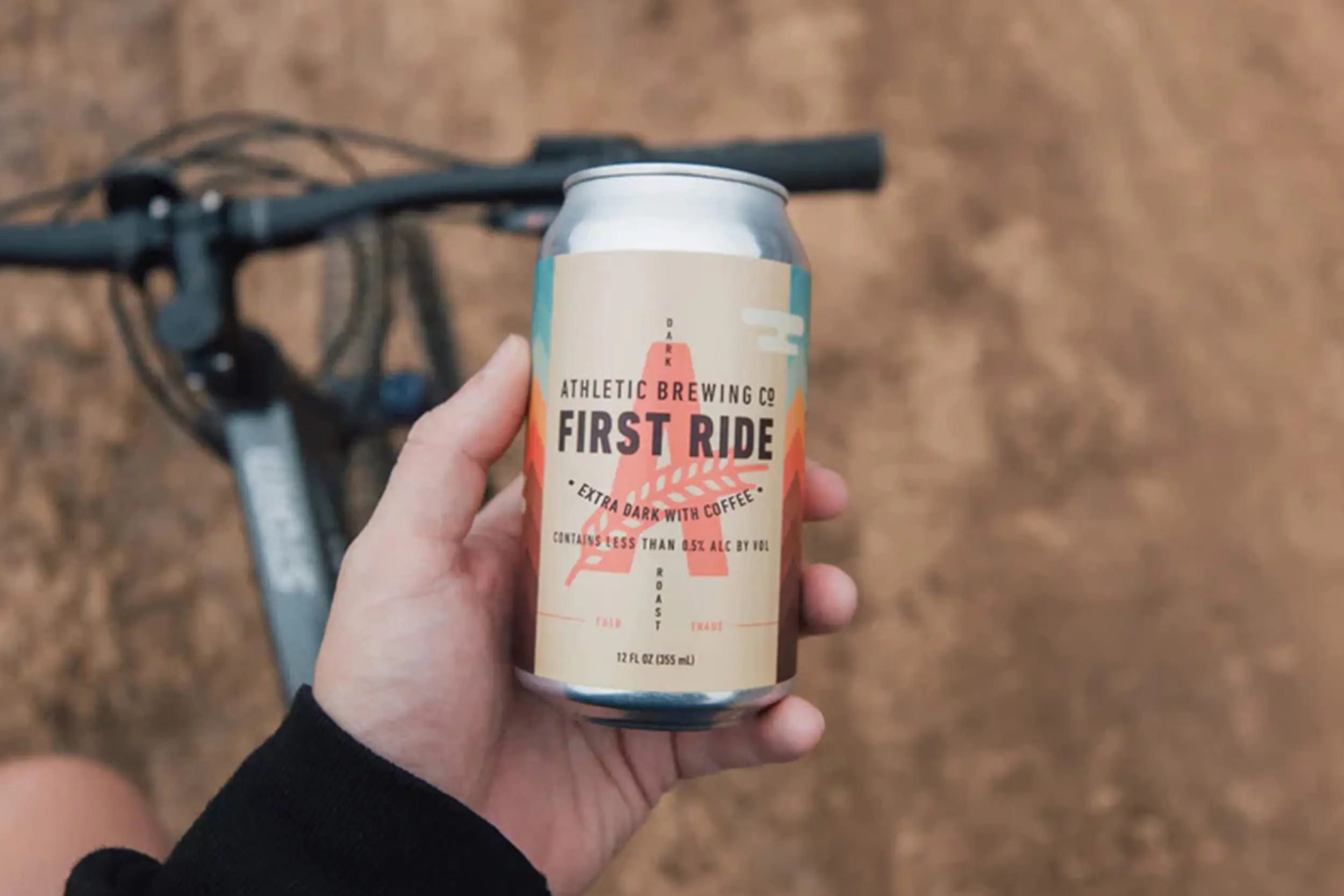 a hand holding a can of athletic brewing company beer with a bicycle in the background