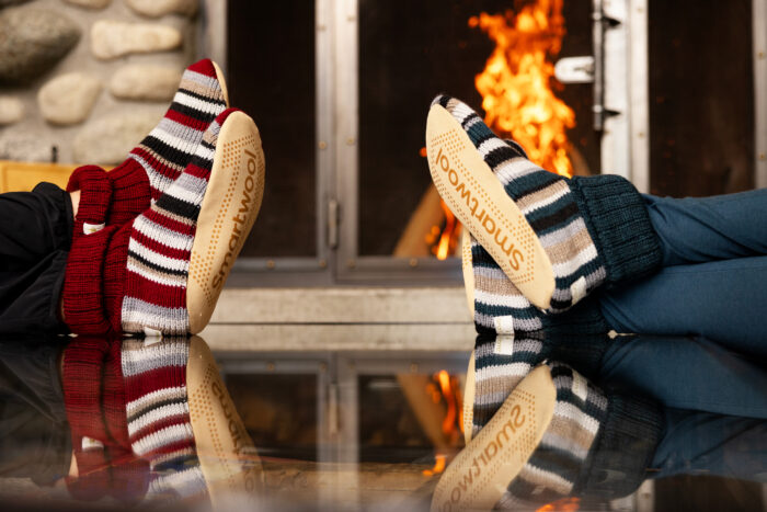 two pairs of legs with smartwool hudson slippers in front of a fireplace