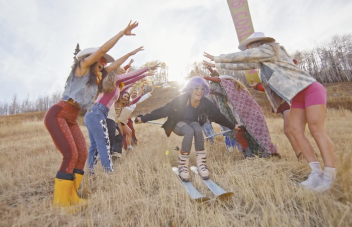 a group of female skiers dress in cowboy boots and flannels as a women with pink hair flies through a tunnel on grass skis