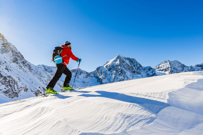 Mountaineer backcountry ski walking up along a snowy ridge with skis in the backpack. In background blue sky and shiny sun and Zebru, Ortler in South Tirol, Italy.