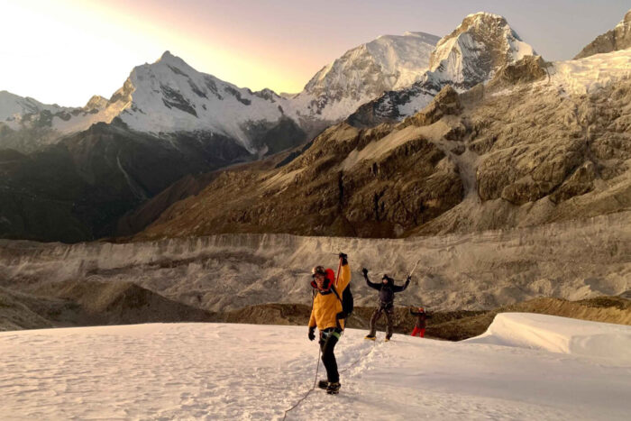 three people raise their hands to the camera while traversing a snowfield with mountains in the background