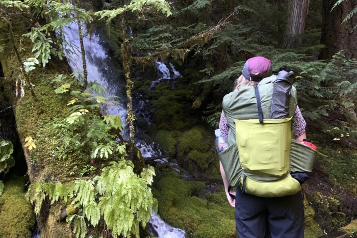 a hiker testing the muted green Palante ultralight pack amongst dense green foliage and waterfall