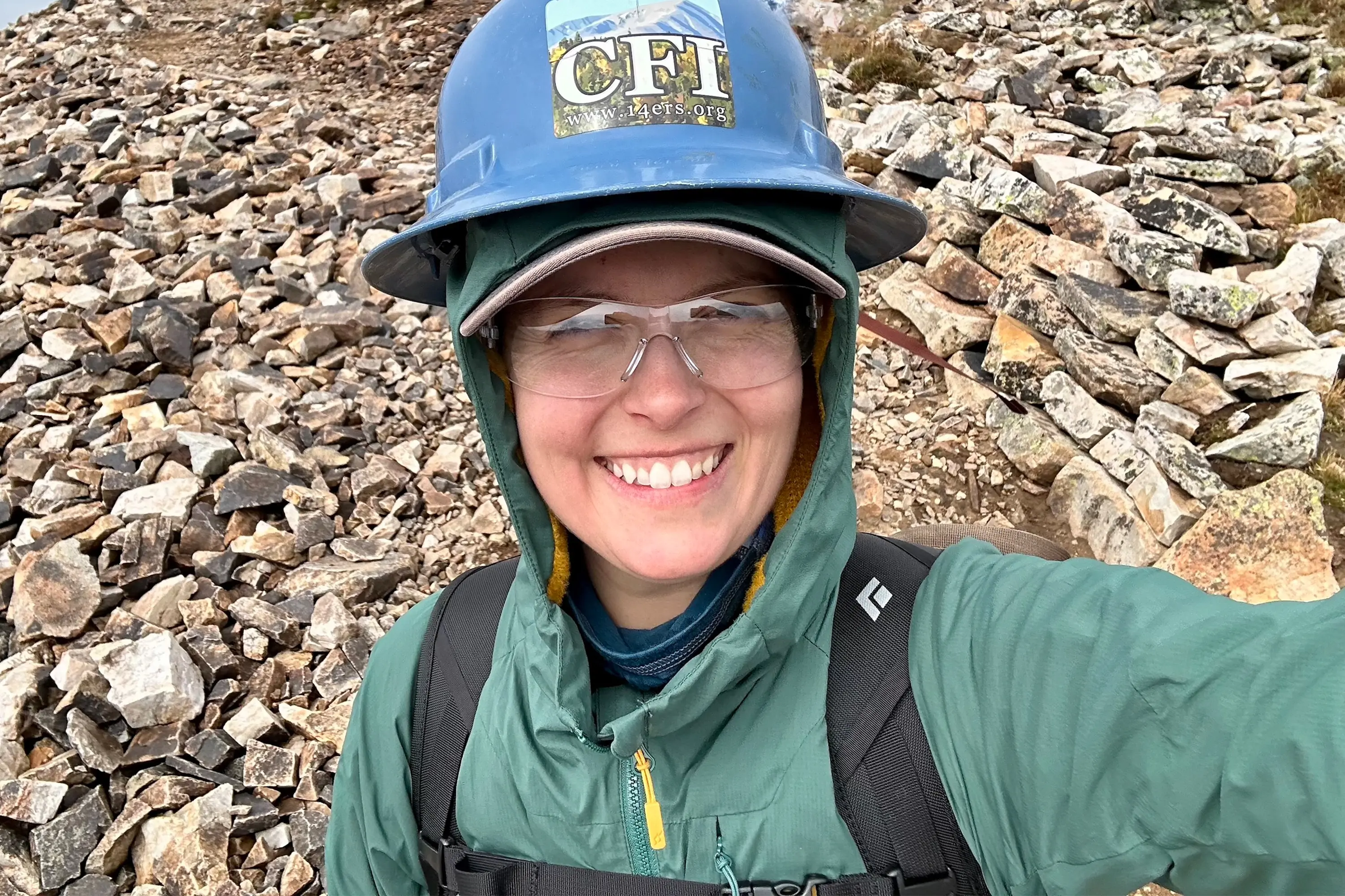 testing the Rab VaporRise Summit Jacket while wearing a work crew helmet in a rock field on a high-alpine peak