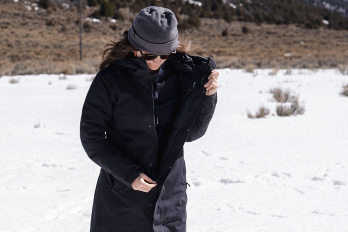 A woman buttoning up her Peter patera parka, in cold windy weathe