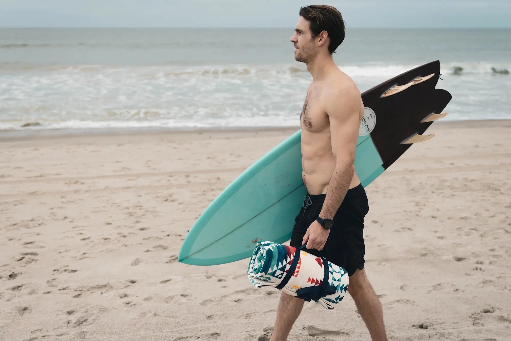 A man carrying the Pendleton Towel for Two and a surfboard to the beach