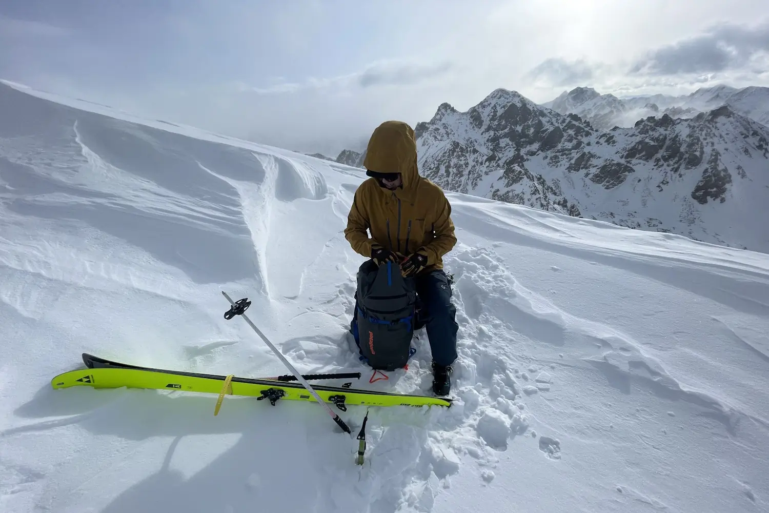 Skier resting on a snowy mountain with gear and snow-covered peaks in the background