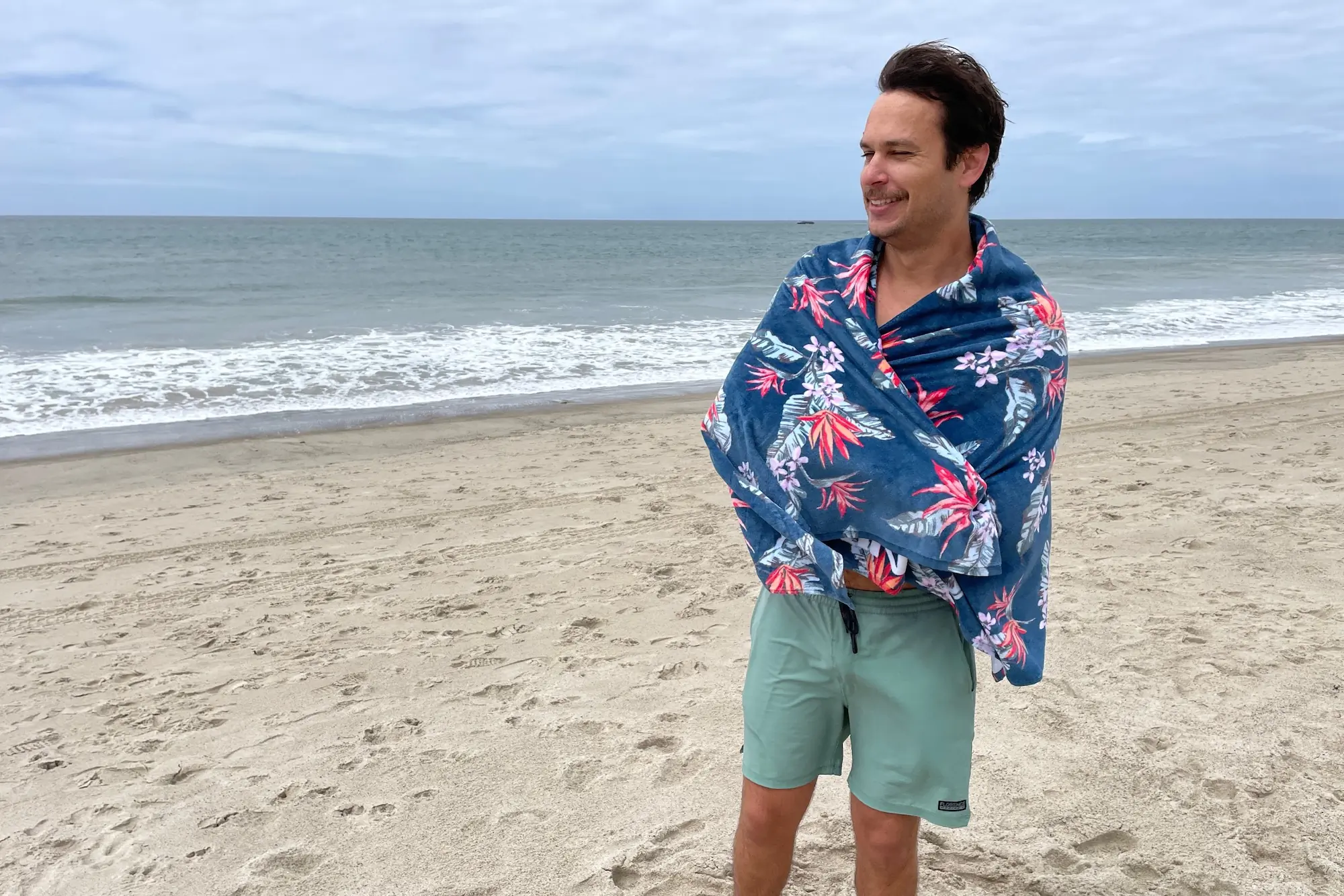 A man wrapped in the Dakine Terry Beach Towel for warmth on a breezy day at the beach