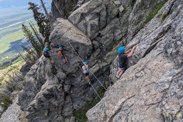 Four climbers in helmets on a rocky via ferrata route above a green valley.