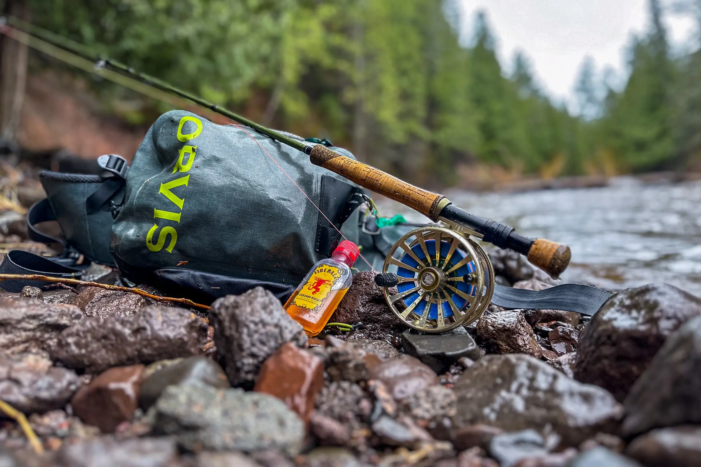 Fly fishing gear on a rocky riverbank with an Orvis backpack, rod, reel, and a Fireball whiskey bottle