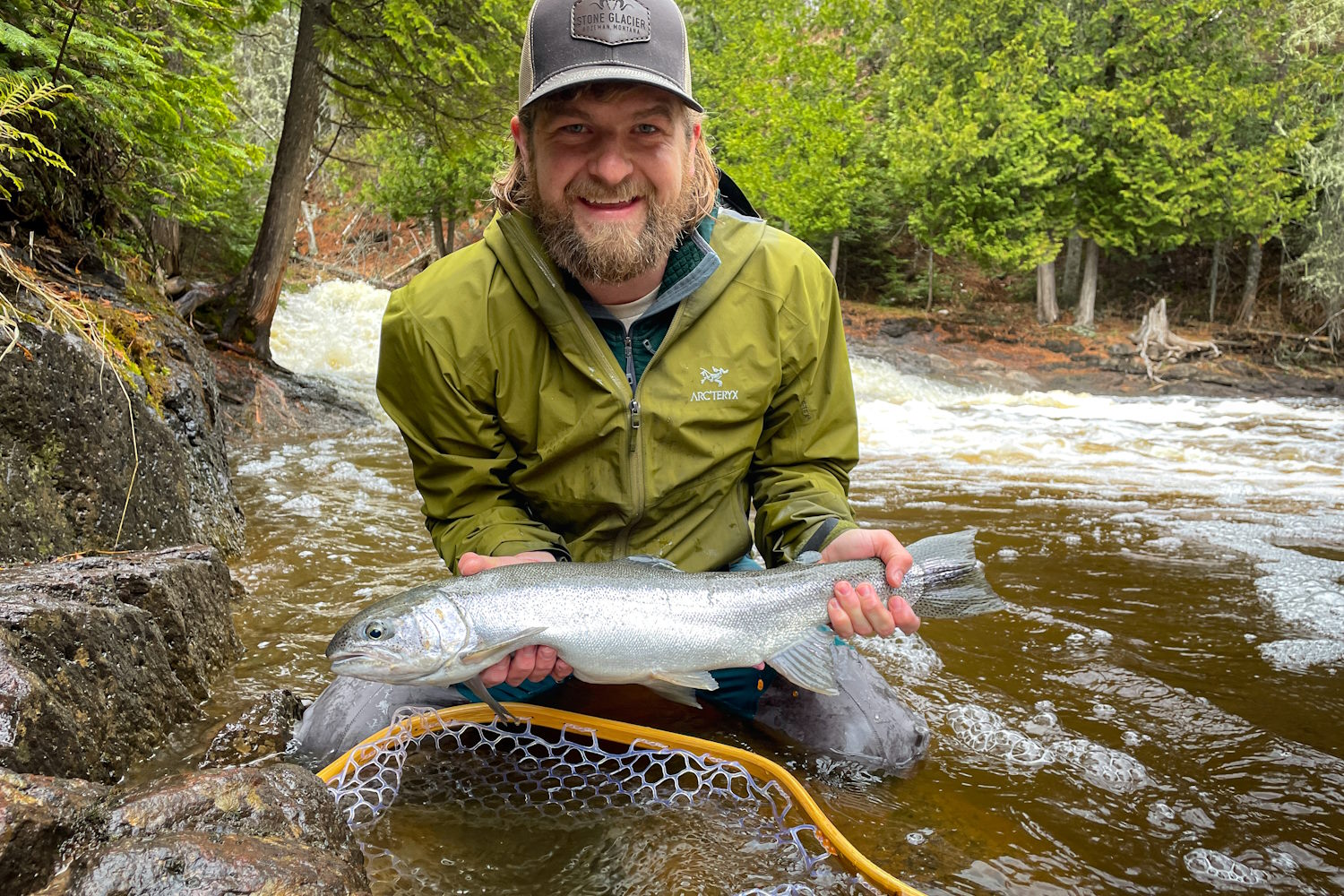 Man holding a freshly caught fish by a stream in a forested area
