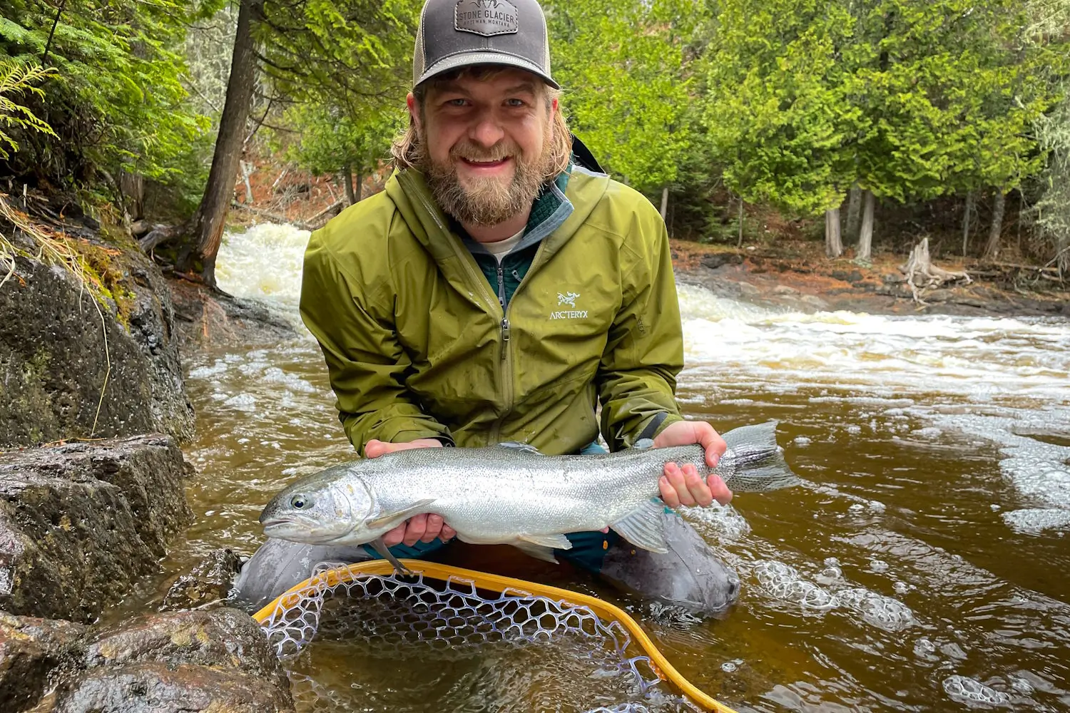 Man holding a freshly caught fish by a stream in a forested area