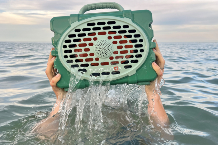 A person is holding a green speaker with both hands above water. Water is streaming out of the speaker.