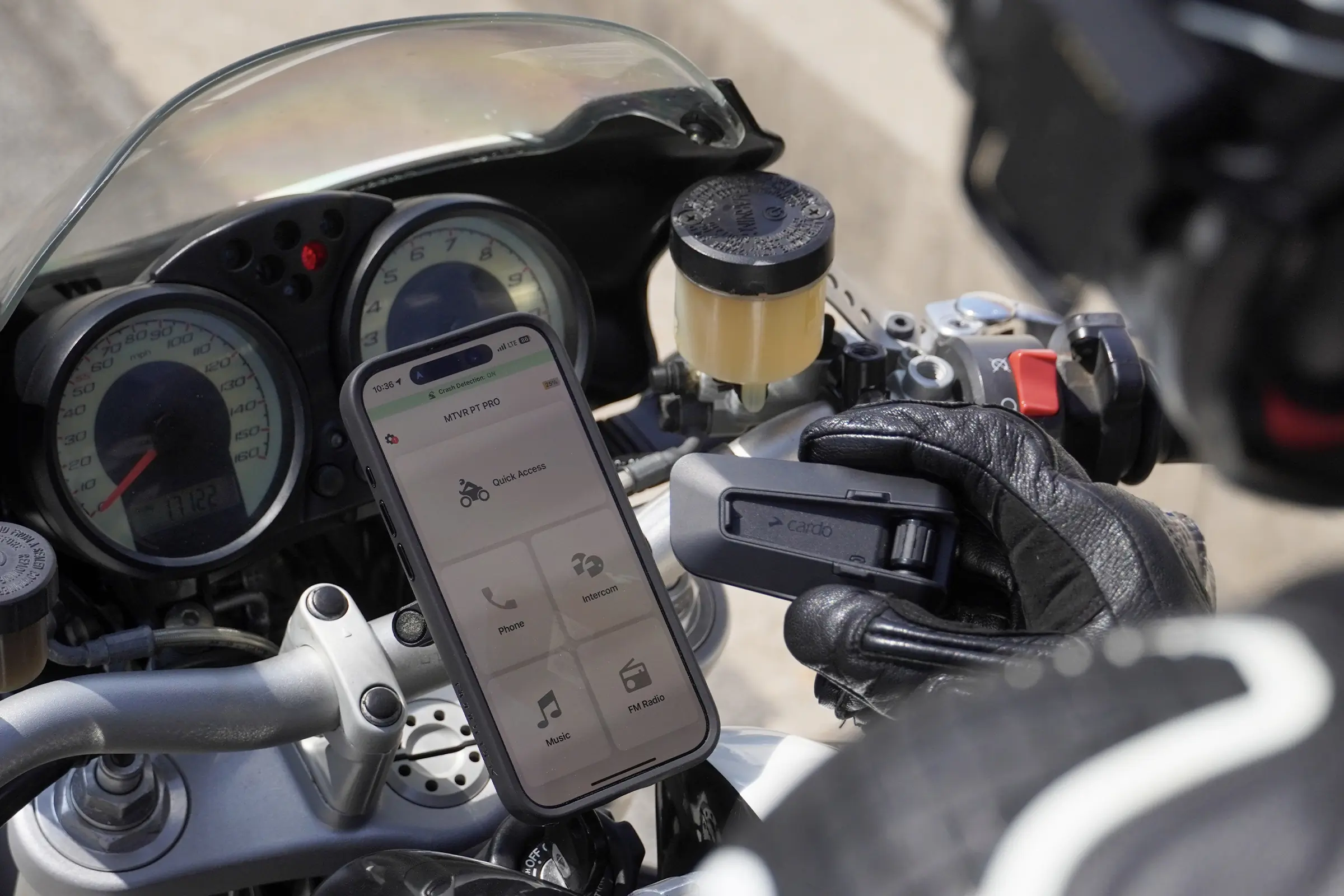 A motorcyclist holds a phone displaying the app and Cardo Packtalk Pro device