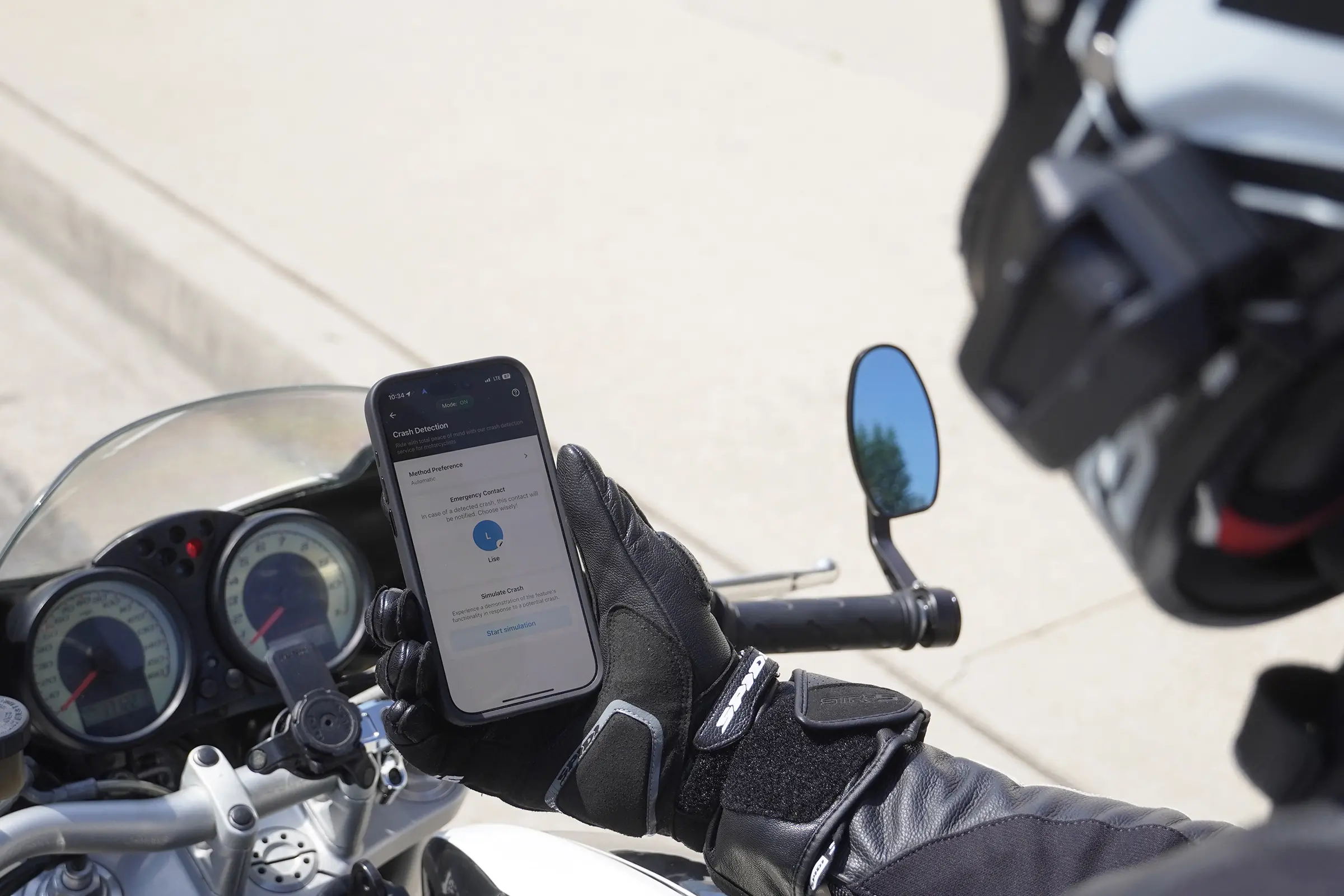 A motorcyclist holds a phone on the motorcycle, displaying the app with crash detection on the screen
