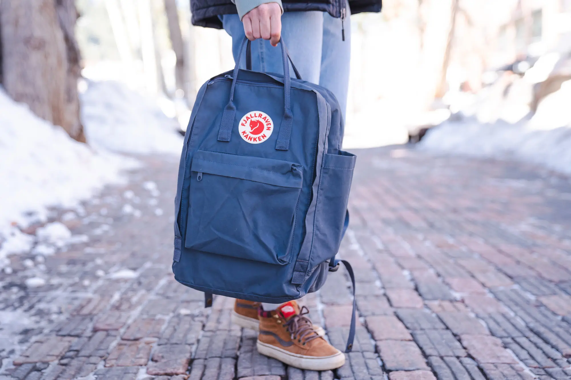 The fjallraven kanken pack being held by the top handles on a snowy sidewalk