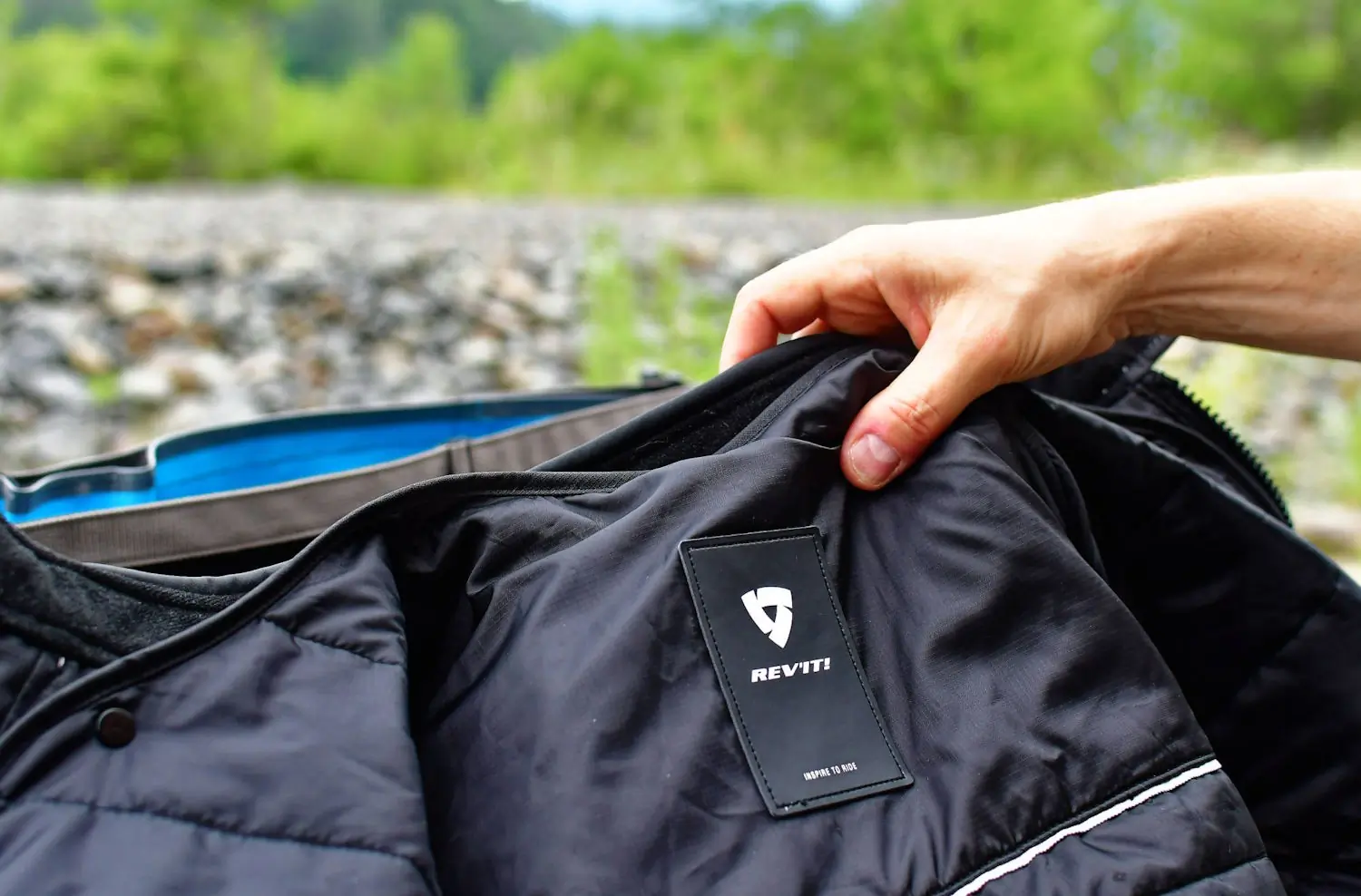 A person holding open a black REV'IT! jacket, showing the inside label with the brand logo