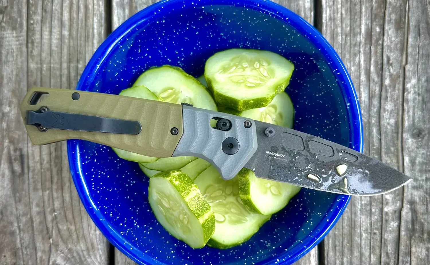 Benchmade PSK knife slicing fresh cucumbers in a blue speckled bowl, placed on a wooden surface