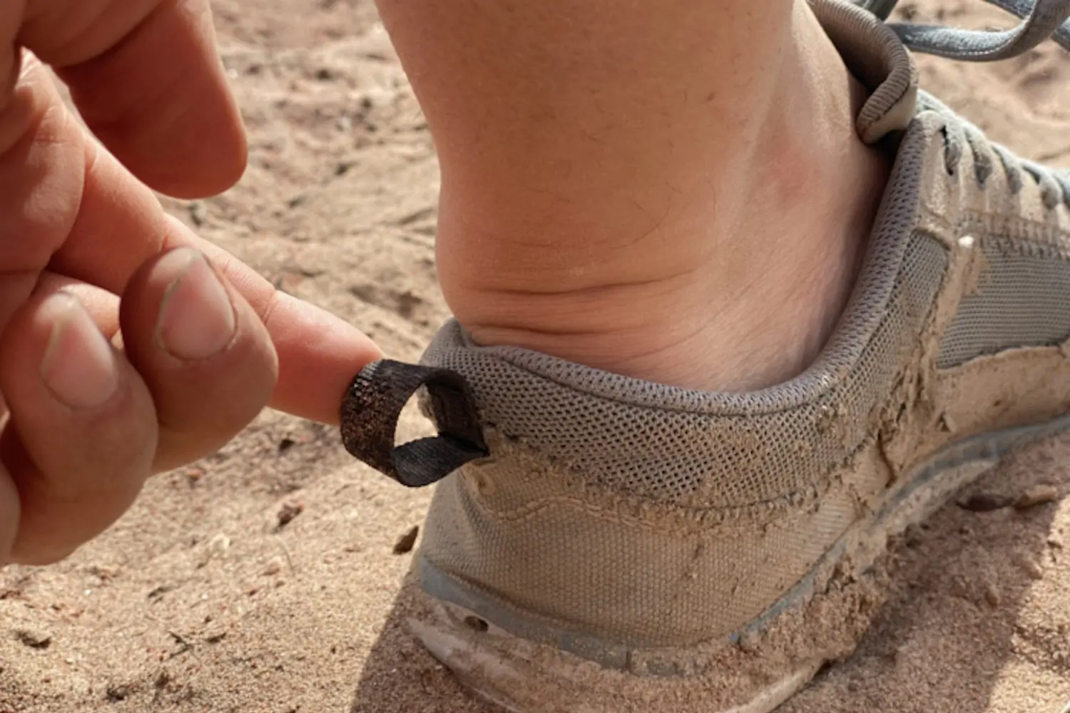 A person pulling the loop on the back of a sand-covered shoe while standing on the sandy ground