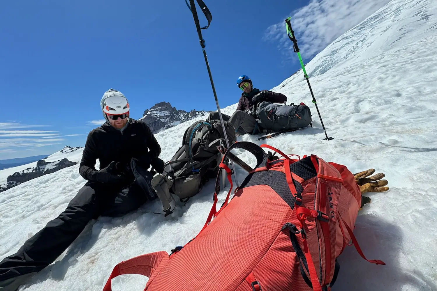 Two climbers take a break on a snowy mountain slope, their gear and Blue Ice backpacks resting on the snow