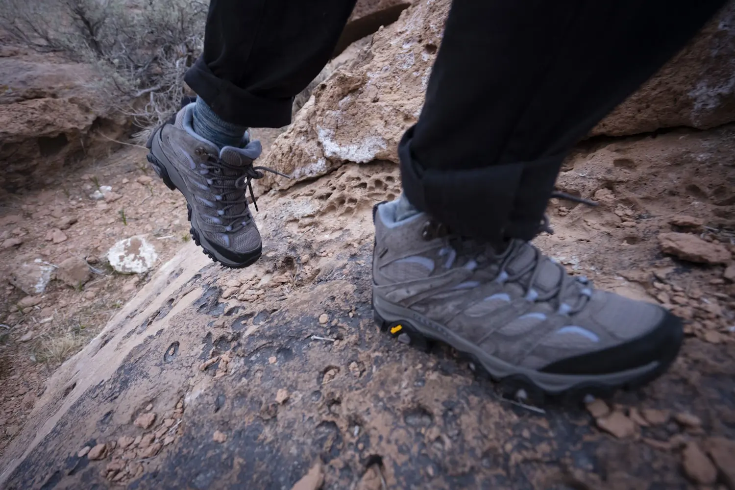 A close-up of a person hiking on rocky terrain, wearing gray moab hiking boots and black pants