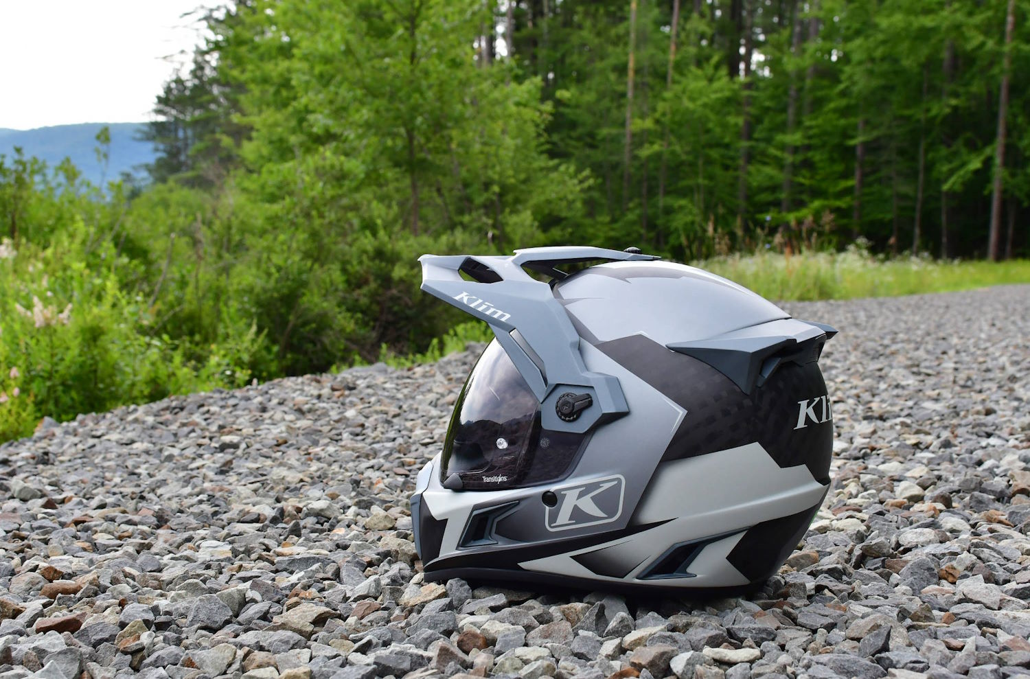 A grey and black motorcycle helmet rests on gravel with a backdrop of lush green trees and bushes