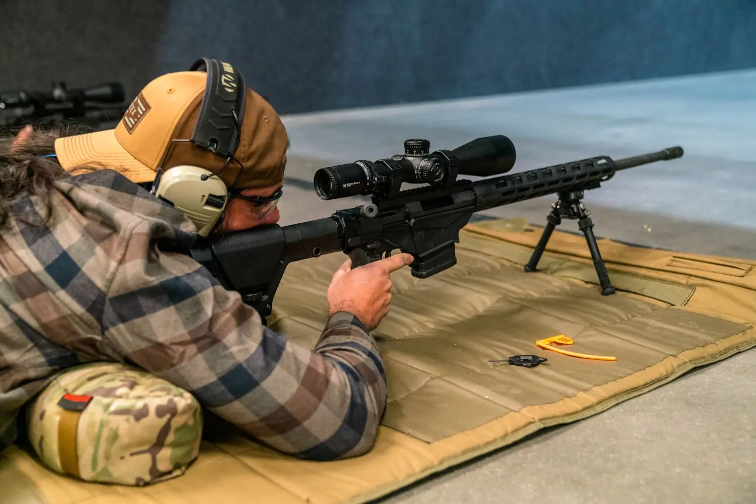 A man lying prone on a mat, aiming a rifle with a scope at an indoor shooting range