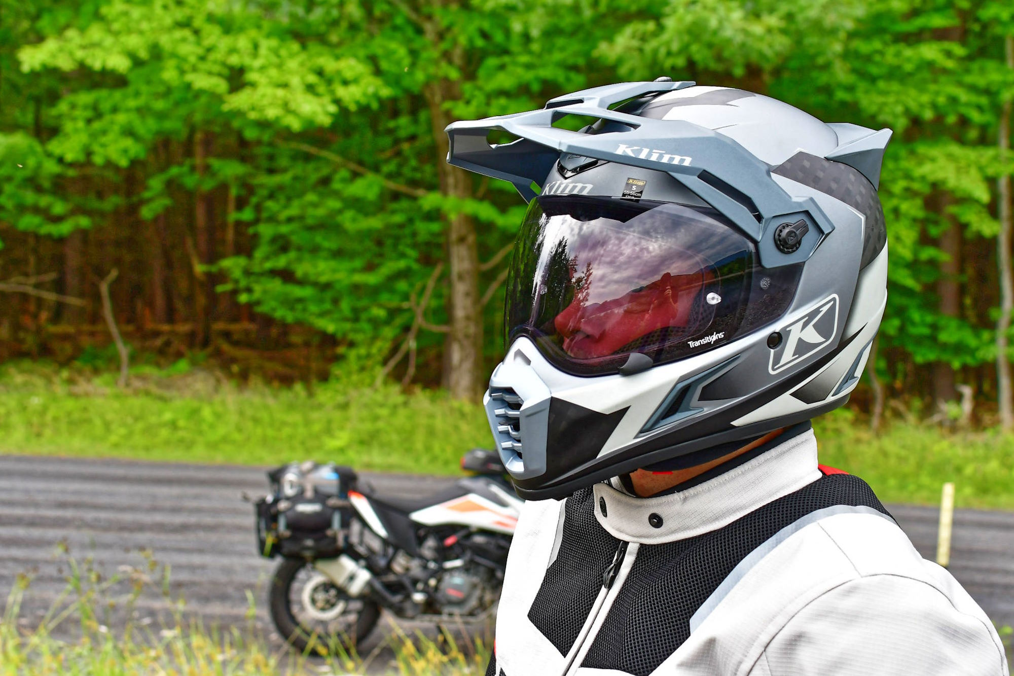 A motorcyclist wearing a Klim helmet stands beside a parked motorcycle on a forest road