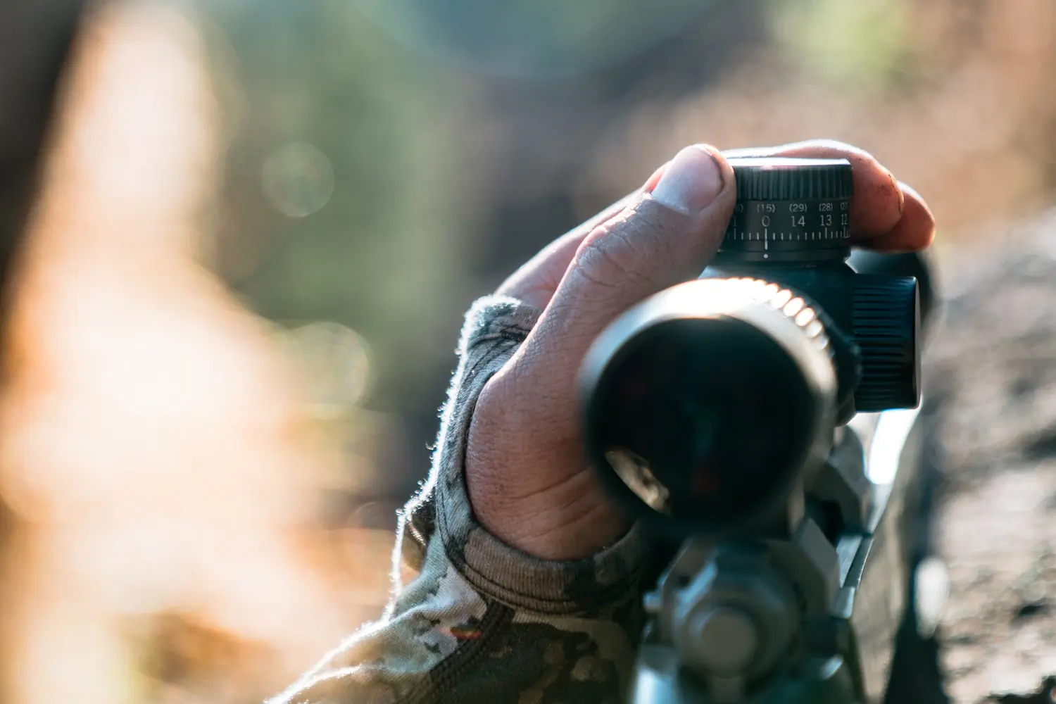 A person adjusts the dial on a rifle scope, wearing camouflage sleeves