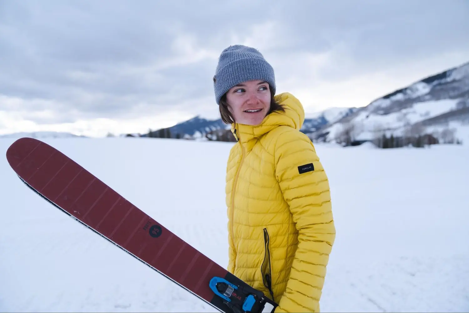 A person in a yellow Decathlon Forclaz MT100 Hooded Down Puffer Jacket holding skis in a snowy mountain landscape on a cloudy day