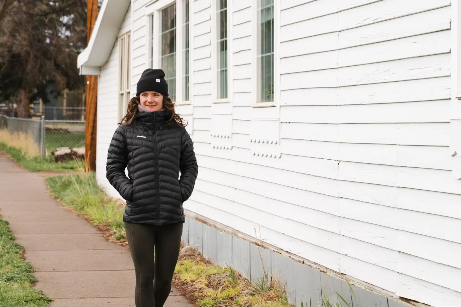 A person in a black Mammut jacket and beanie walks along a sidewalk next to a white wooden building