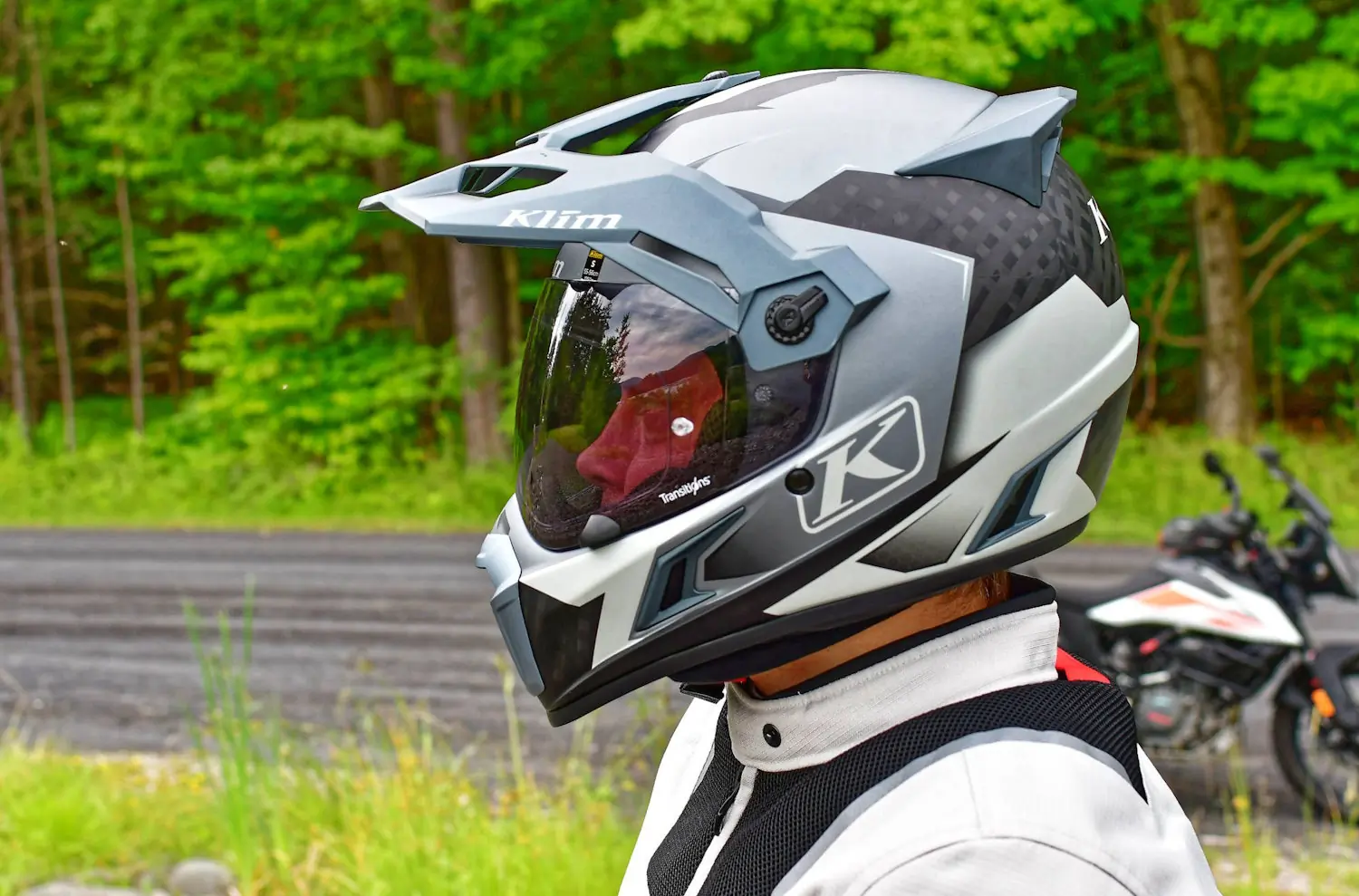 A person wearing a grey Klim motorcycle helmet stands near a road with a motorcycle in the background, surrounded by green trees