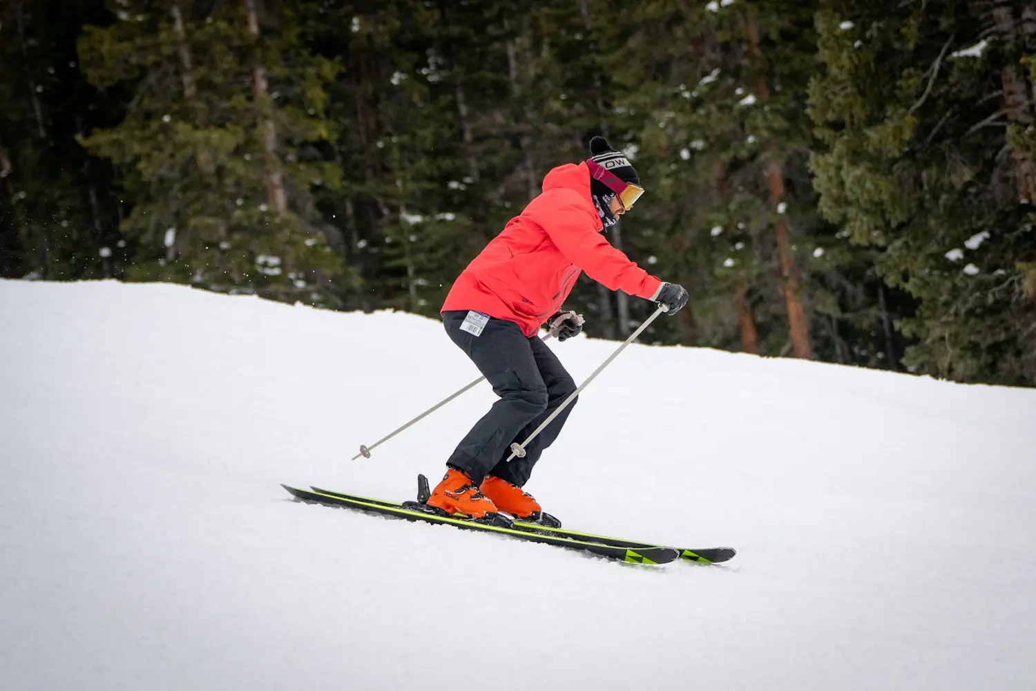 Skier in a red jacket and orange boots skiing downhill on Fischer skis, holding ski poles for balance