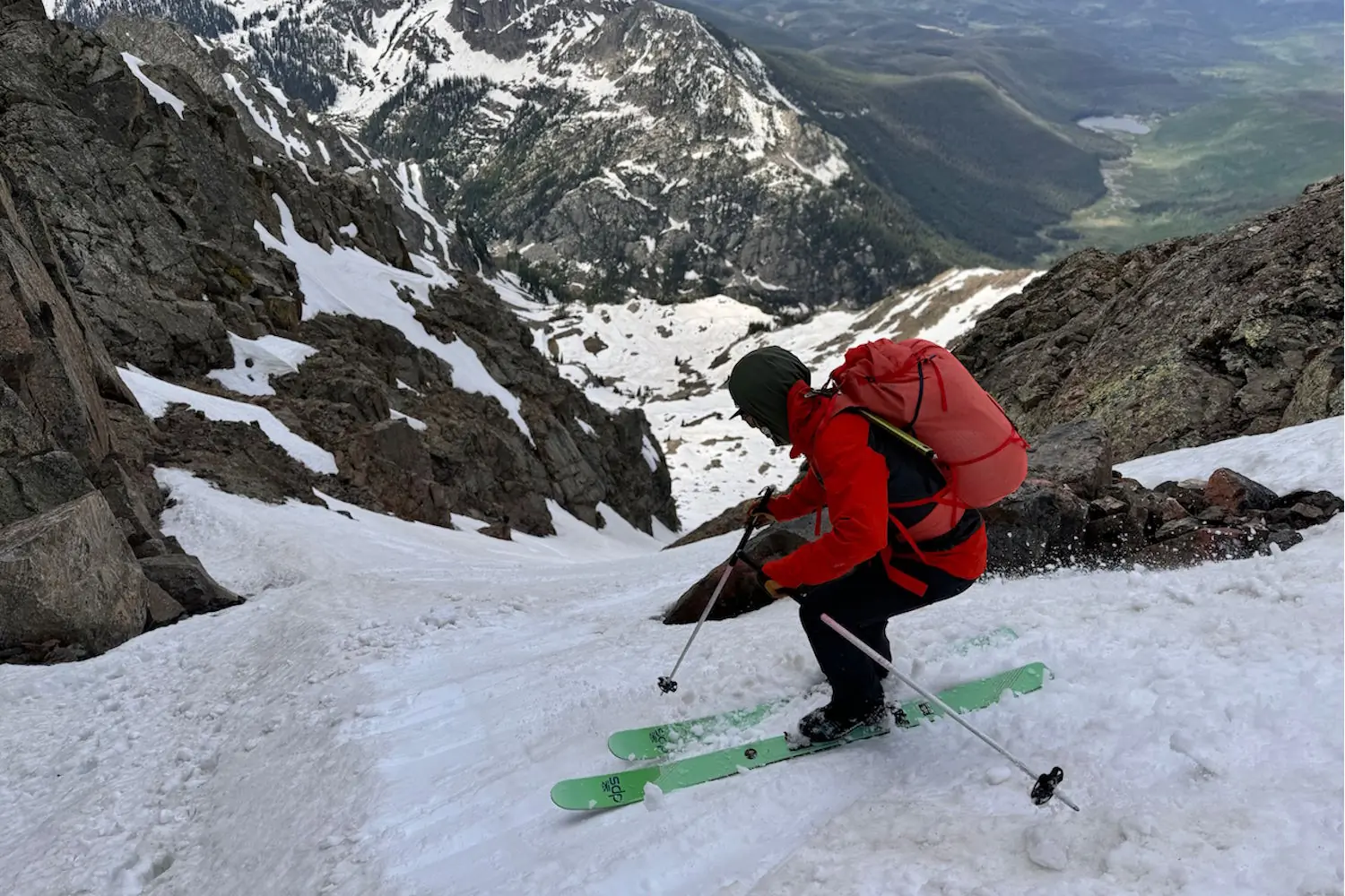 Skier with a red backpack navigating a steep, snow-covered mountain slope with rocky terrain 