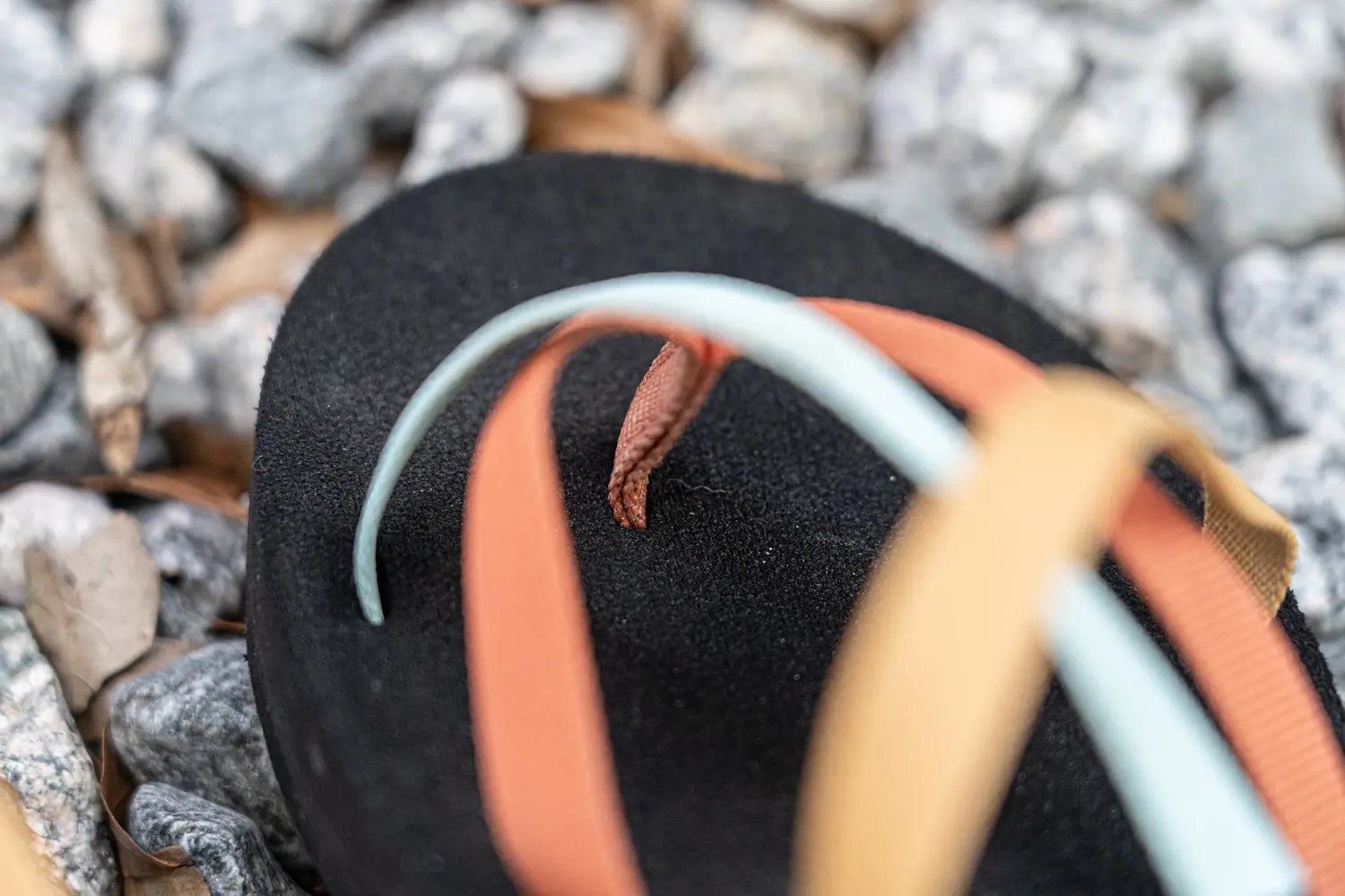 A close-up of a teva olawahu sandal with crisscrossing straps in light blue, orange, and beige, on a rocky surface