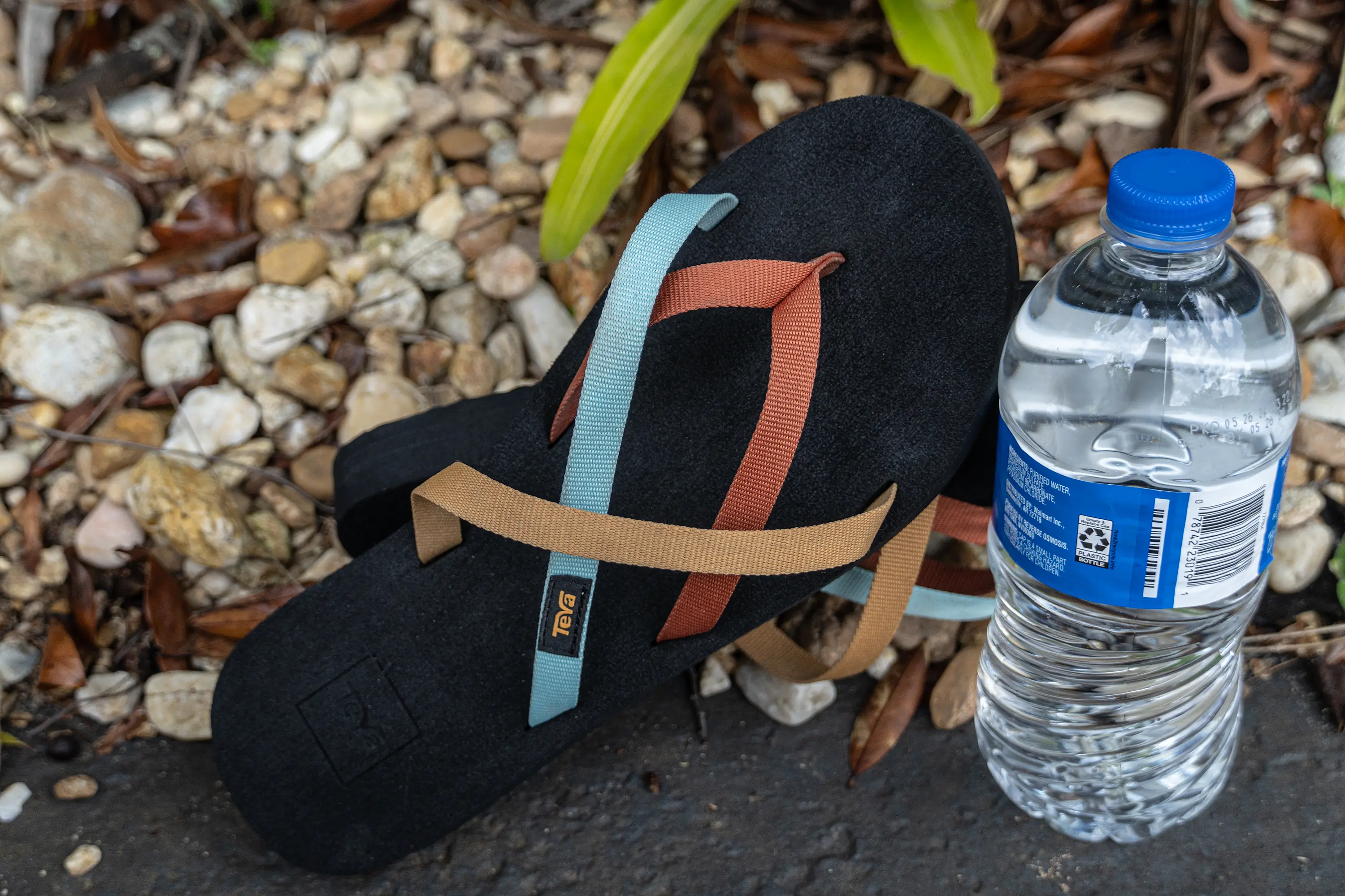 A pair of black Teva Olowahu with multicolored straps next to a plastic water bottle, placed on a rocky surface