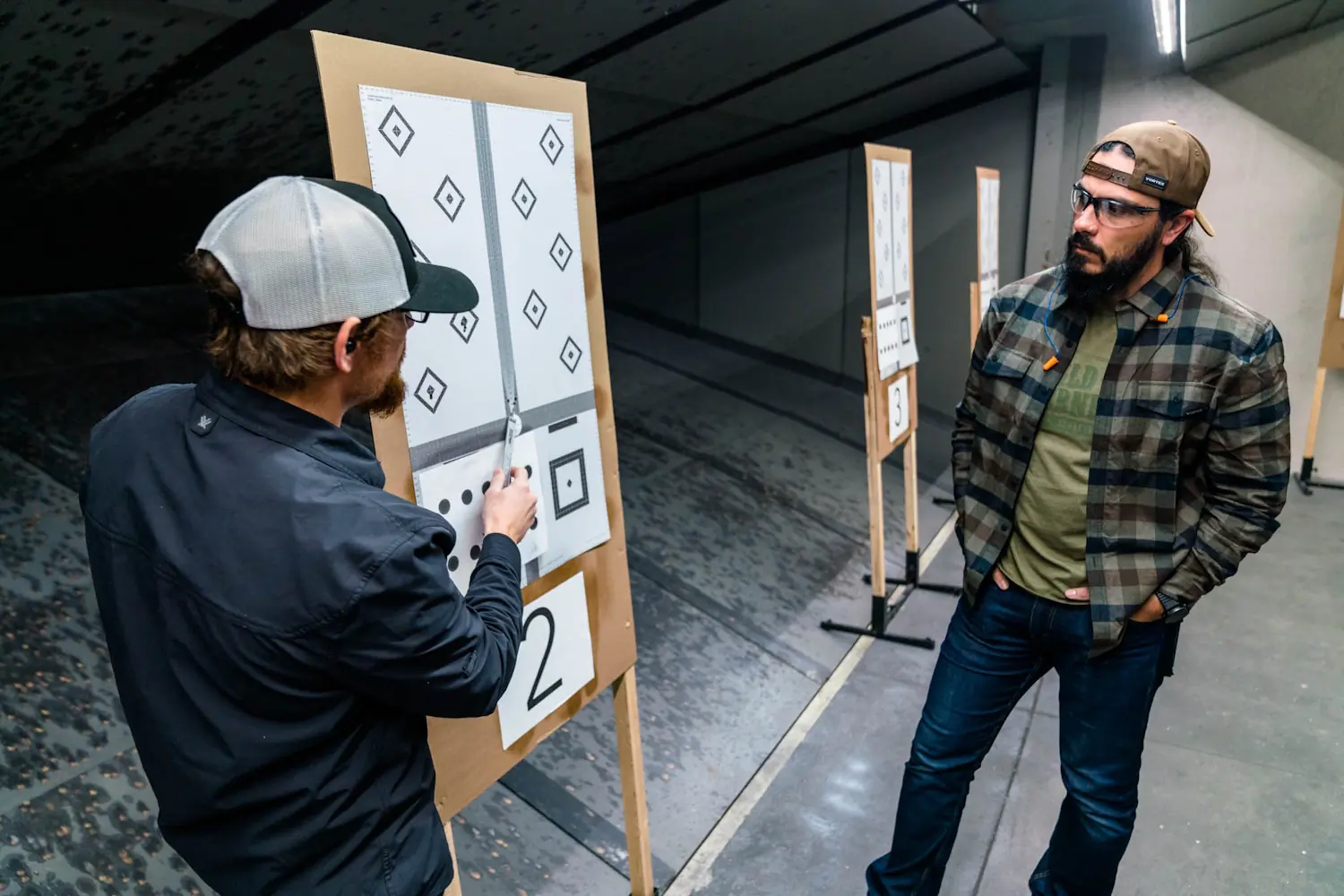 Two men at an indoor shooting range examining targets; one man is pointing at a target with a tool, while the other man stands nearby observing